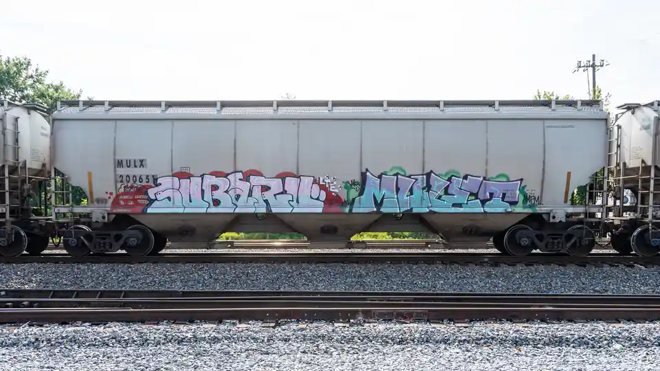 A side view of a white freight train car on tracks, covered in colorful graffiti that reads 'SUBURB MIX'. The car has…