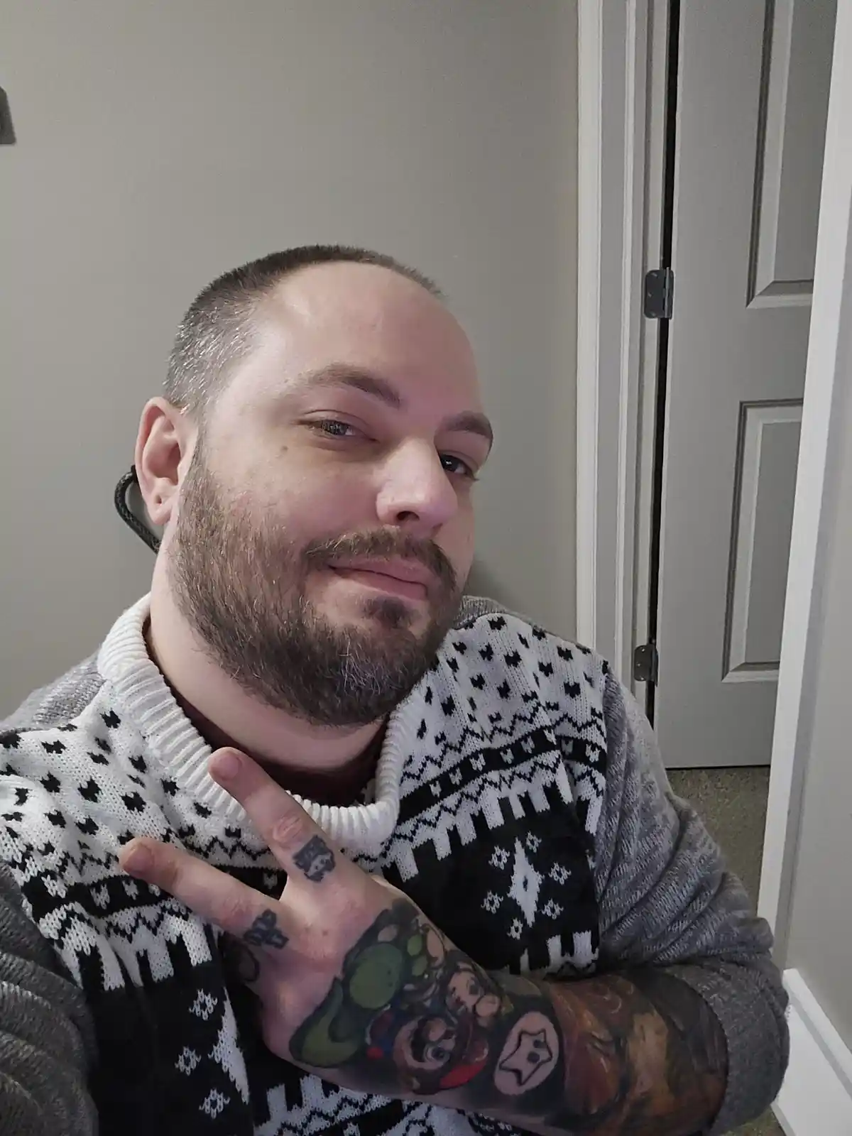 A man with a beard and tattoos on his arm and fingers poses with a peace sign while wearing a black and white patterned…