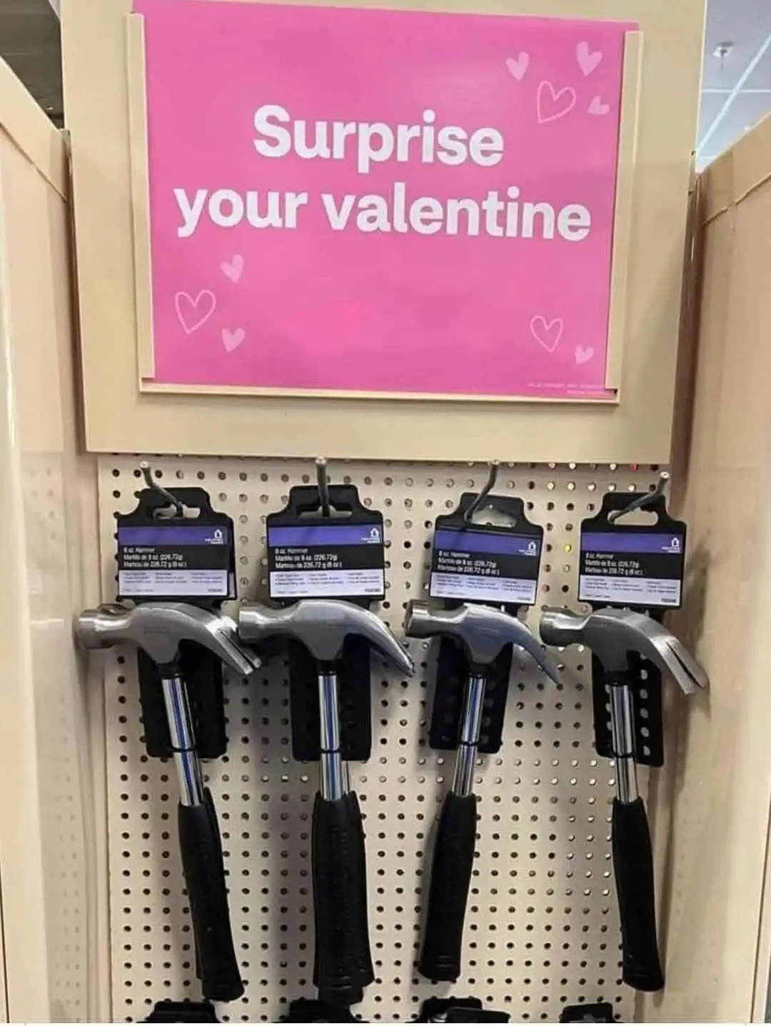 A retail display shows four hammers hanging on a pegboard beneath a pink sign that reads 'Surprise your valentine' with…