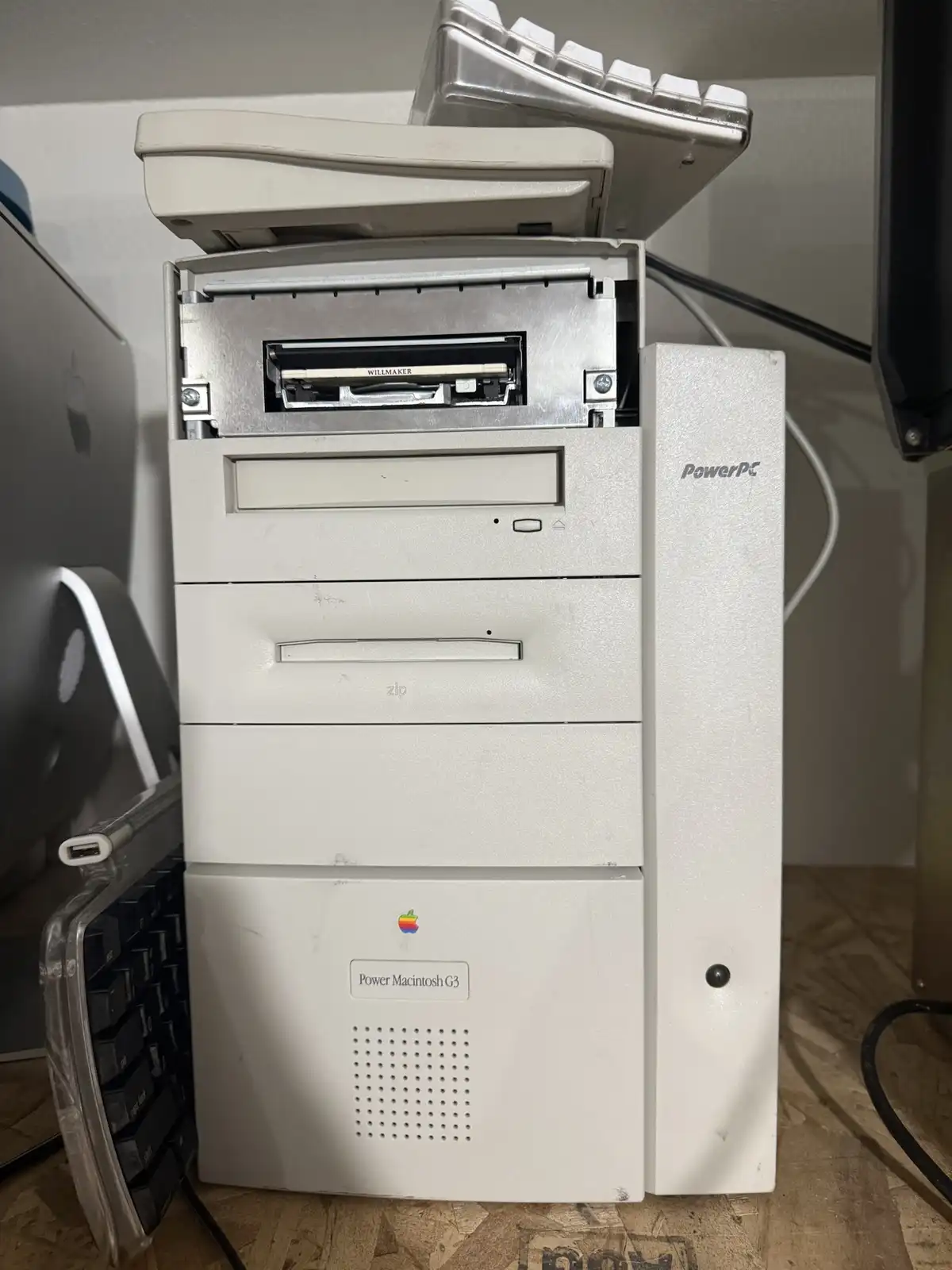 A white Apple Power Macintosh G3 tower computer with its top panel open, revealing an internal drive bay. A silver keyboard…