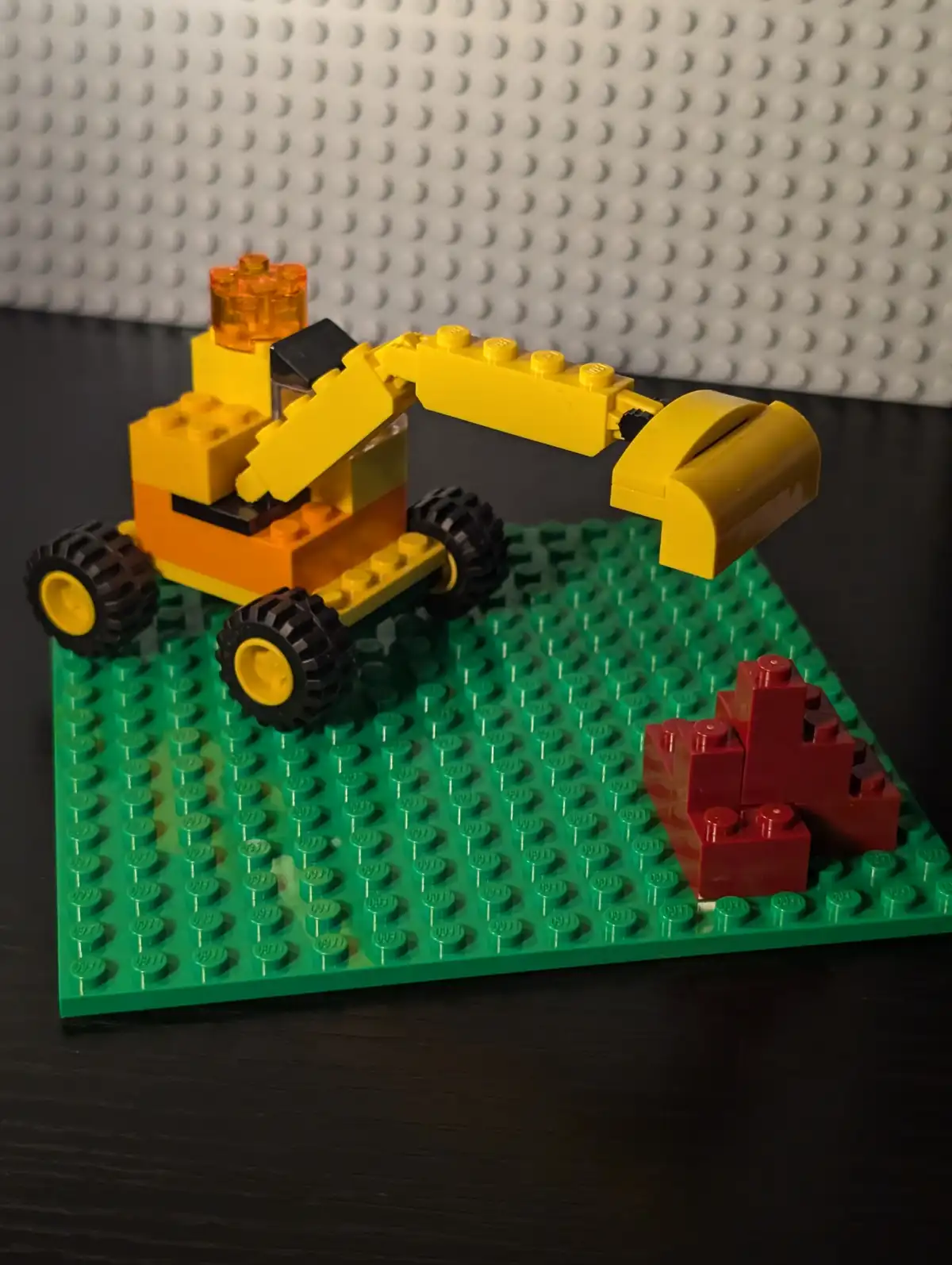 A yellow and orange LEGO excavator with black tires sits on a green LEGO baseplate, its arm extended toward a small pile of…