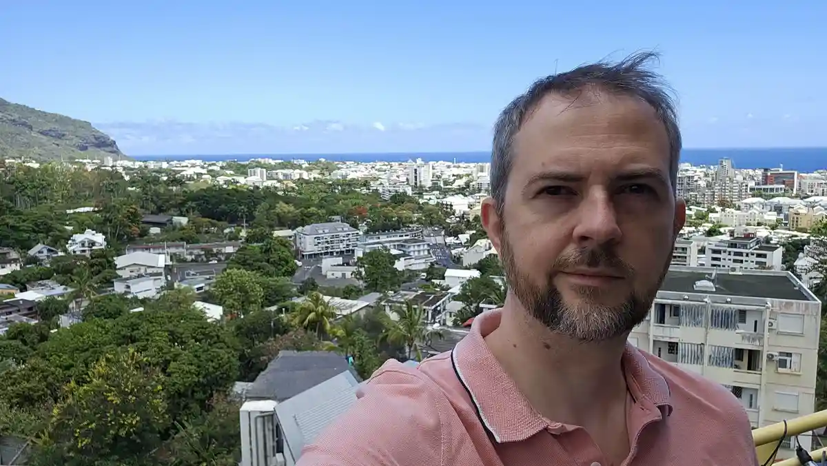 A man with a beard and pink polo shirt takes a selfie overlooking a scenic coastal city with mountains and ocean in the…