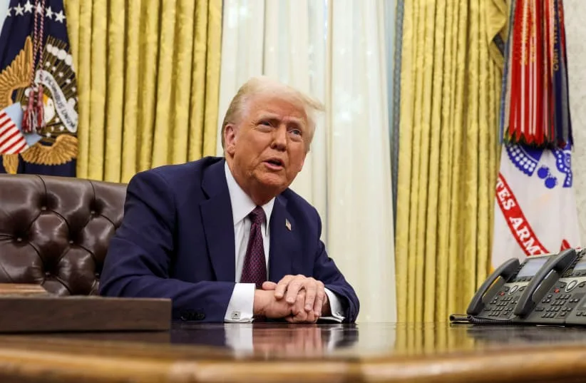 a man in a dark suit and tie sits at a large wooden desk in an official-looking room with gold curtains and presidential…