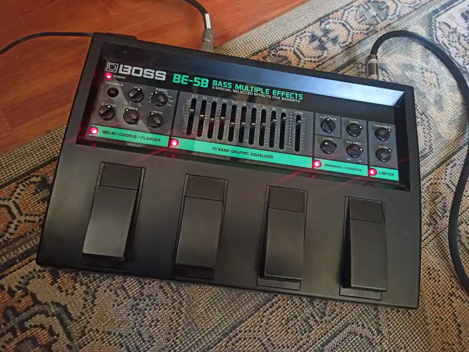 A black Boss BE-5B bass multi-effects pedal with red power and effect indicator lights is shown on a patterned rug. The…