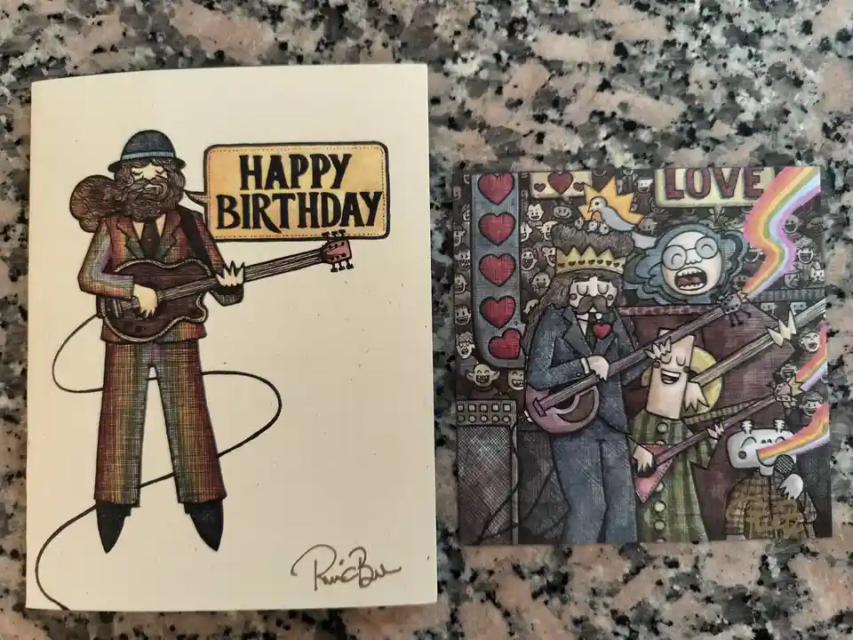 Two illustrated birthday cards by artist R. B. are shown side-by-side on a granite countertop. The left card features a…