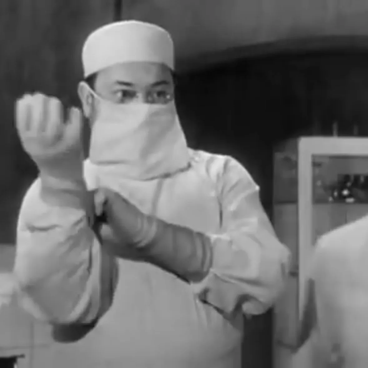 A black-and-white image shows a person in surgical attire, including a cap, mask, and gloves, making a gesture with their…