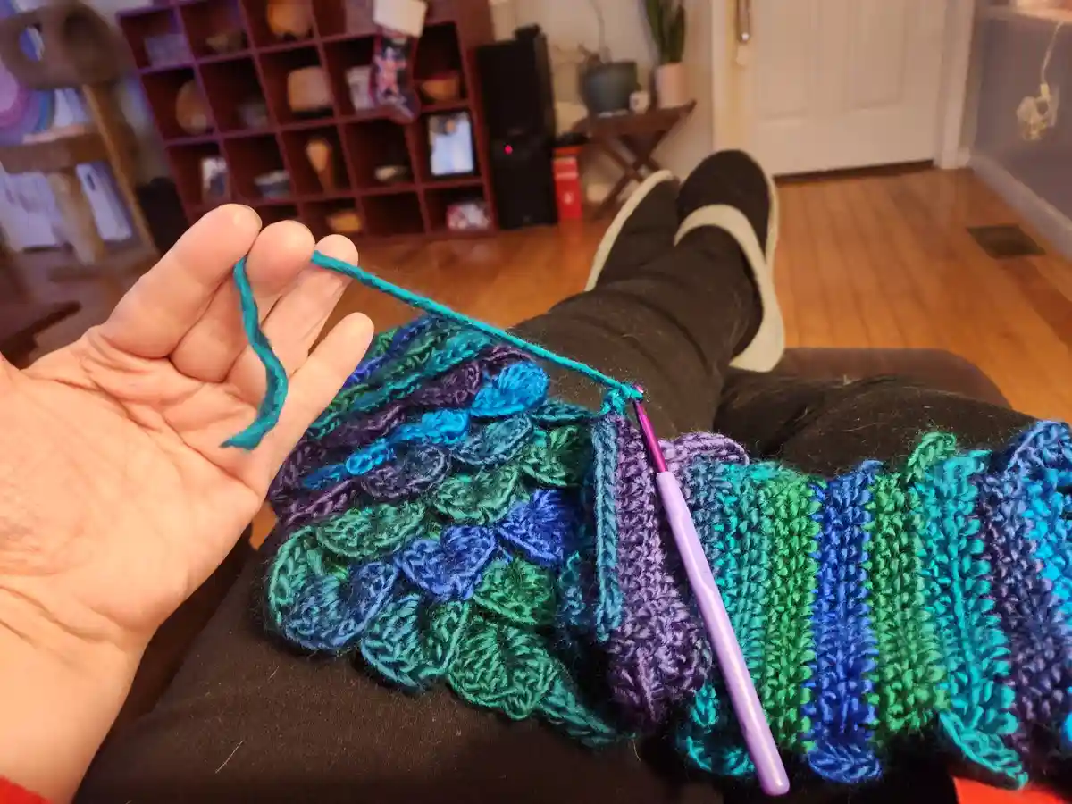A person is lying on their back with a colorful, multicolored crocheted piece resting on their lap, holding the yarn with…