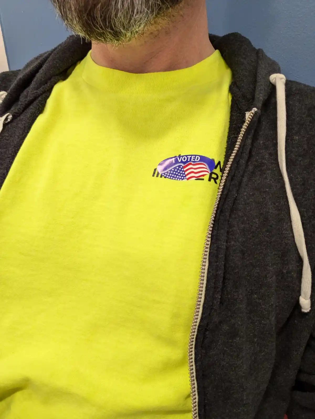 A close-up of a person wearing a bright yellow t-shirt under a dark grey zip-up hoodie, showing a small 'I VOTED' sticker…