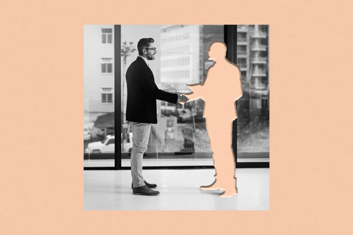 A man in a suit and glasses shakes hands with a solid peach-colored silhouette of another person, both standing indoors near…