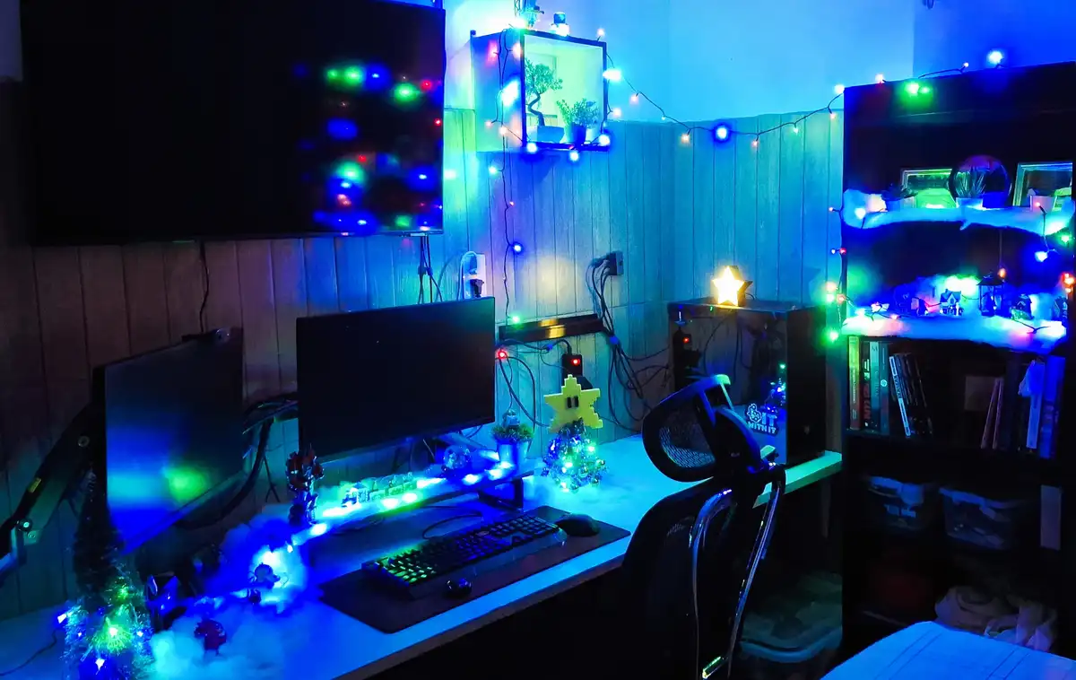A dimly lit computer desk setup is decorated with glowing blue and green string lights, fake snow, miniature Christmas…