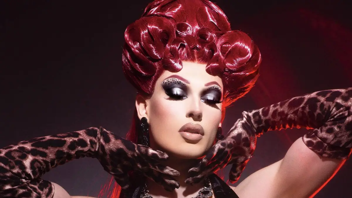 A person in drag with a large, shiny red wig, heavy black and silver eye makeup, and leopard-print gloves frames their face…