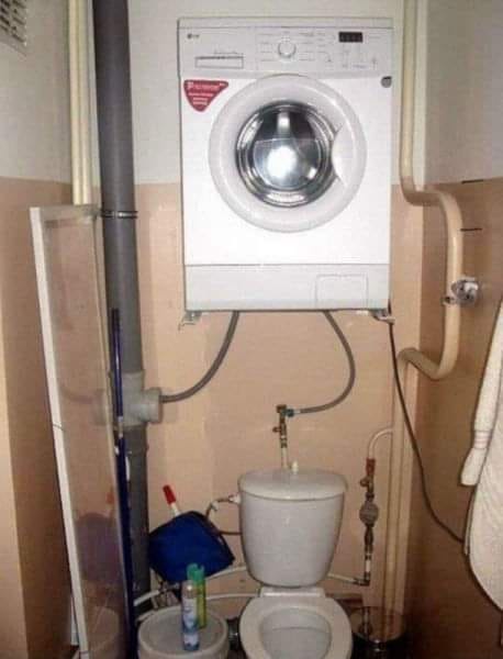 A white front-loading washing machine is mounted directly above a toilet in a cramped bathroom, with pipes and hoses visibly…