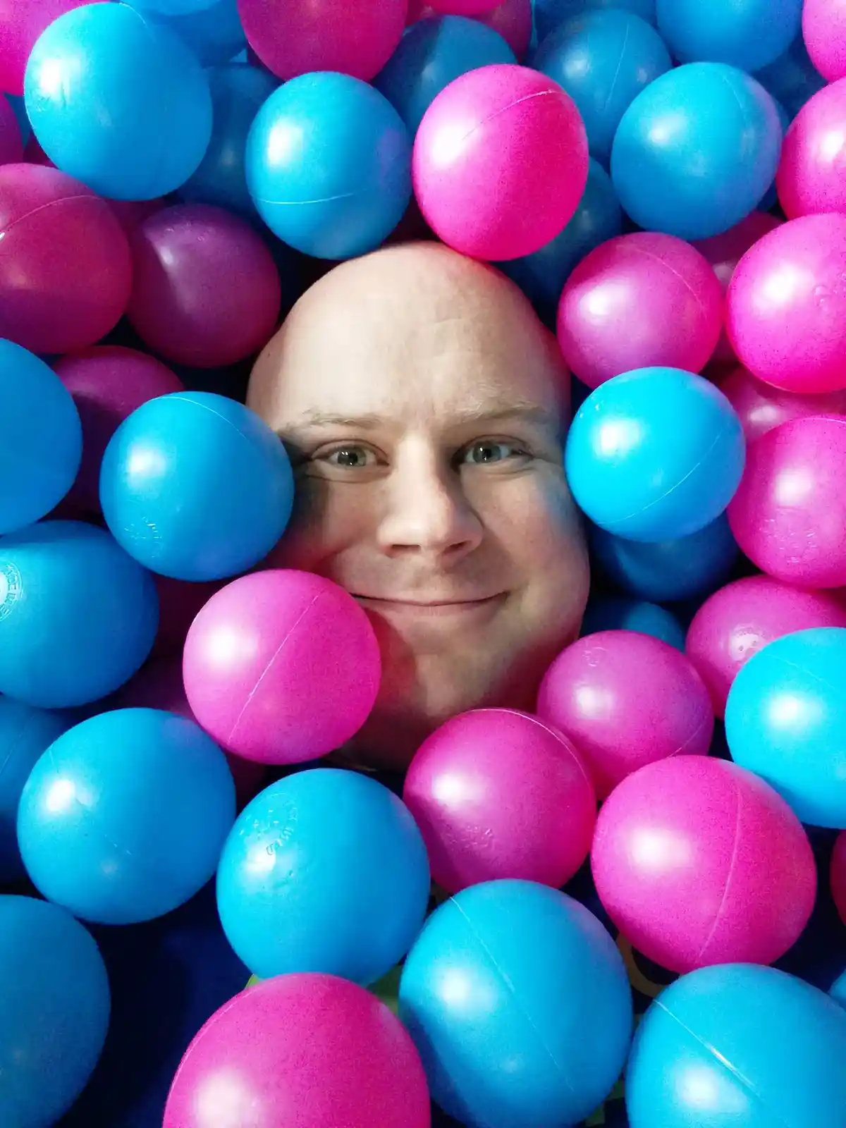 A bald man with a slight smile peers out from a sea of bright blue and hot pink plastic balls, with the text 'i'm not a ball…
