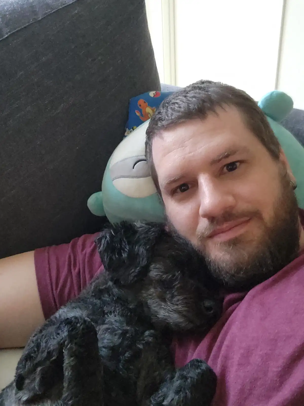A man with a beard and short hair, wearing a maroon t-shirt, takes a selfie while holding a small, fluffy black dog. Behind…
