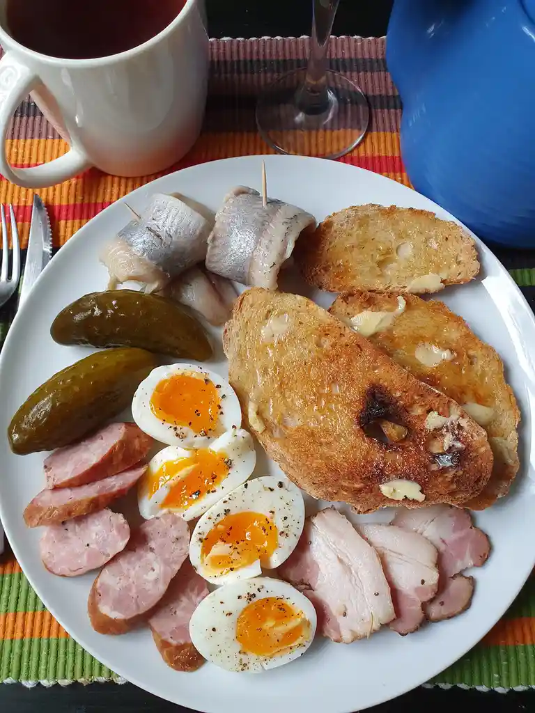 A plate of breakfast food including sliced boiled eggs, ham, sausage, pickles, buttered toast, and rolled fish, with a mug…
