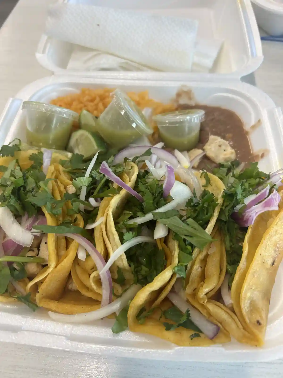 A close-up photo of a white styrofoam takeout container filled with several soft tacos topped with cilantro, sliced red…