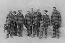 A black-and-white historical photo shows seven men in striped prison uniforms standing in a line outdoors. The image has a…