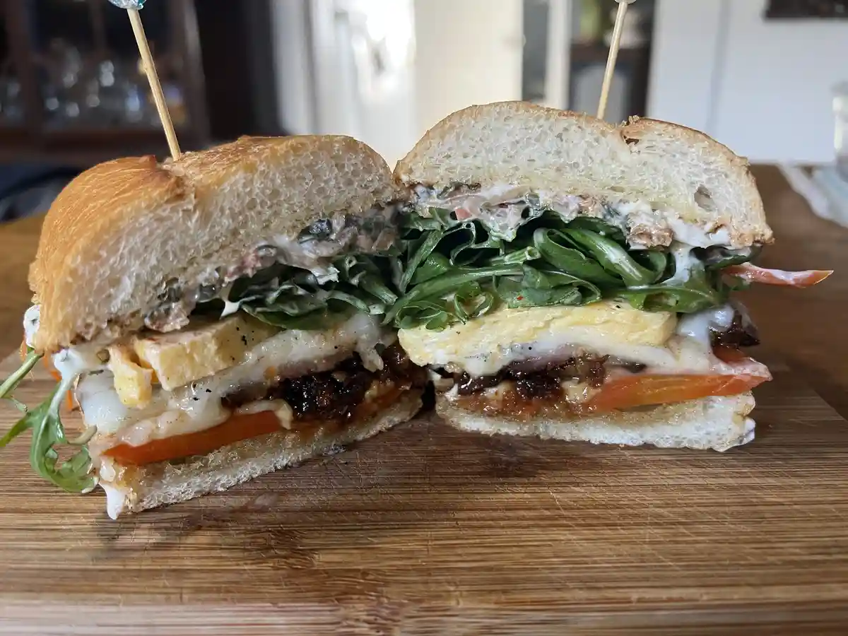 A close-up photo shows a large sandwich cut in half, revealing layers of melted cheese, arugula, tomato, and other…