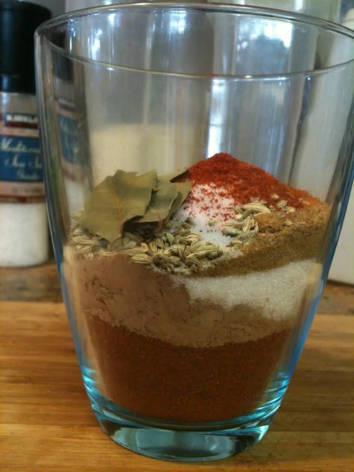 a clear glass tumbler on a wooden surface holds layered spices: a bottom layer of reddish-brown powder, a middle layer of…