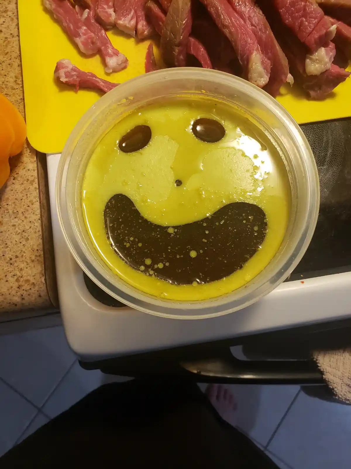 A clear plastic container holds a yellow liquid marinade with two olives as eyes and a dark curved shape as a mouth, forming…