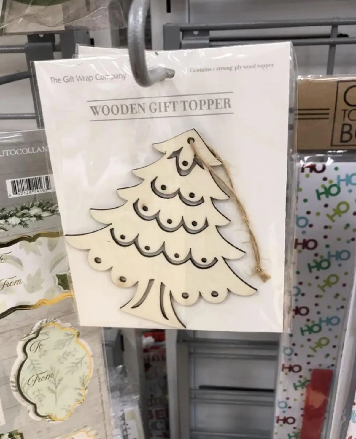 A wooden Christmas tree gift topper from 'The Gift Wrap Company' is displayed in its packaging on a store shelf, featuring a…