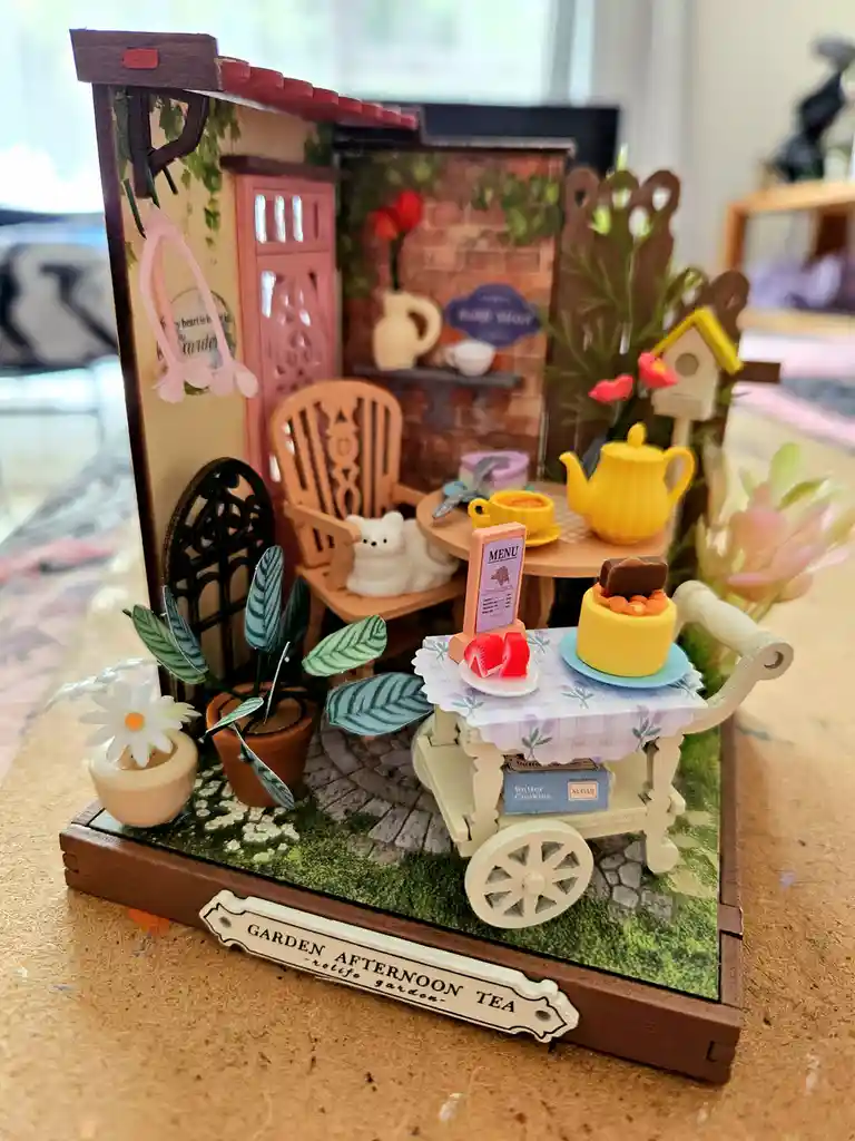 A miniature diorama depicts a charming garden tea set with a tiny white cat curled up in a wooden chair, a yellow teapot, a…