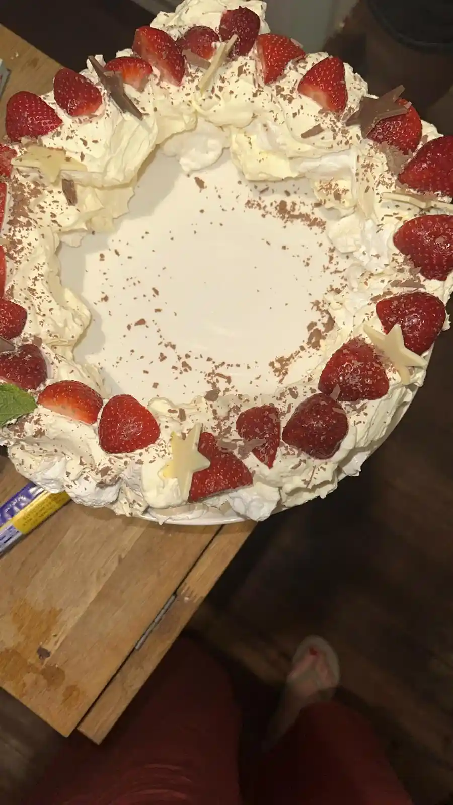 A top-down photo of a cake decorated with whipped cream, sliced strawberries, and chocolate shavings, with the center…