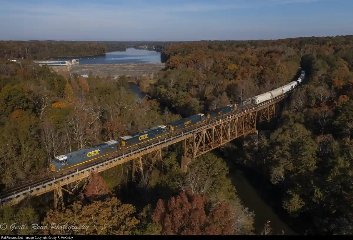 An aerial photograph shows a long CSX freight train crossing a rusty metal trestle bridge over a river, surrounded by dense…
