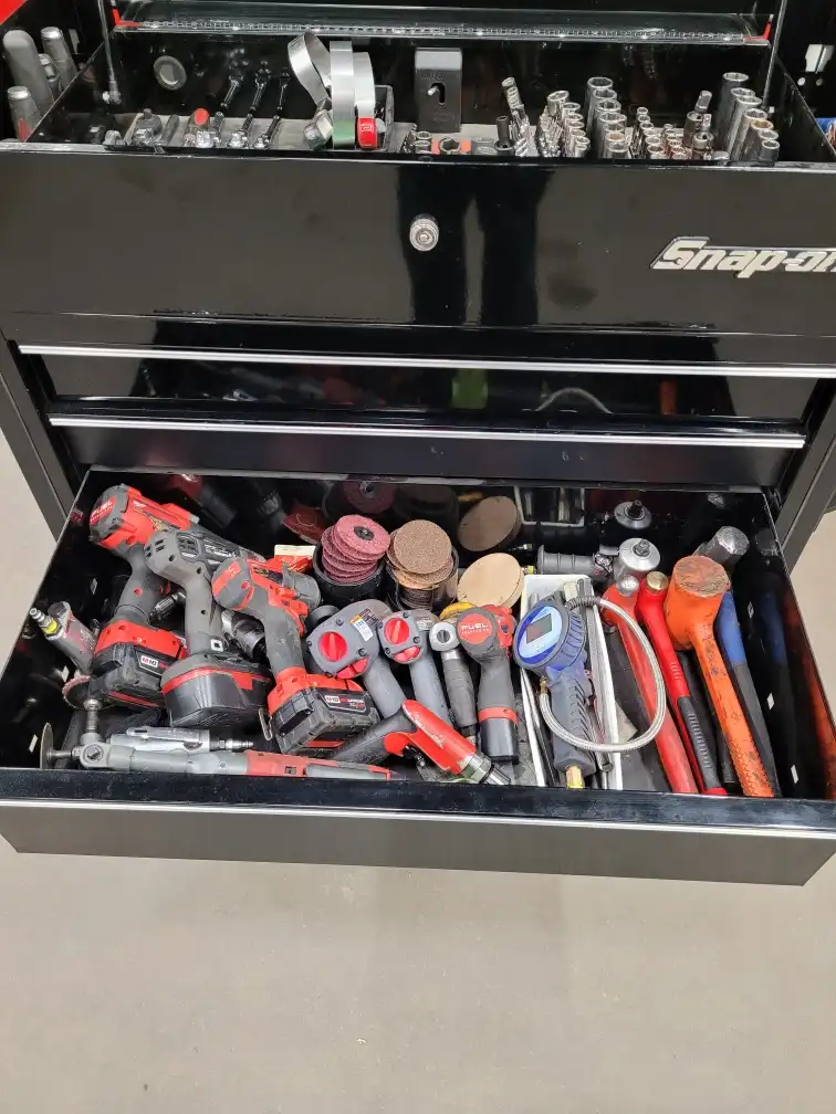 A black Snap-on tool chest with one drawer pulled open, revealing a chaotic pile of power tools, sanding discs, hammers, and…
