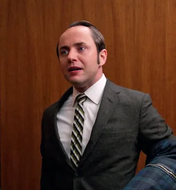 A man in a grey suit and striped tie stands with a surprised, wide-eyed expression, looking upward. He is holding a plaid…