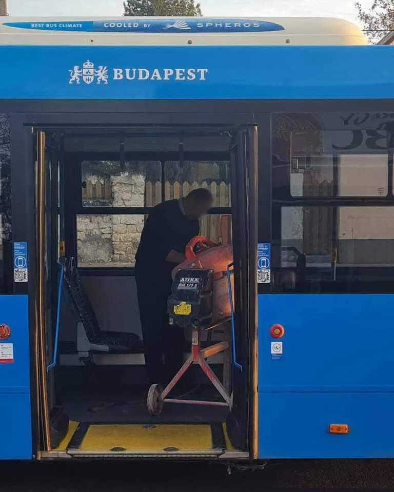 A blue Budapest public bus has its doors open, revealing a man in a dark shirt inside, bent over and operating a large…
