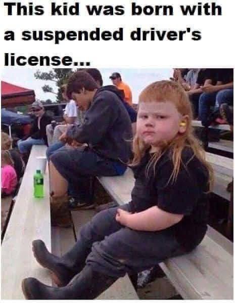 A young child with long red hair and a pouty expression sits on bleachers, wearing a black shirt and dark pants. The…