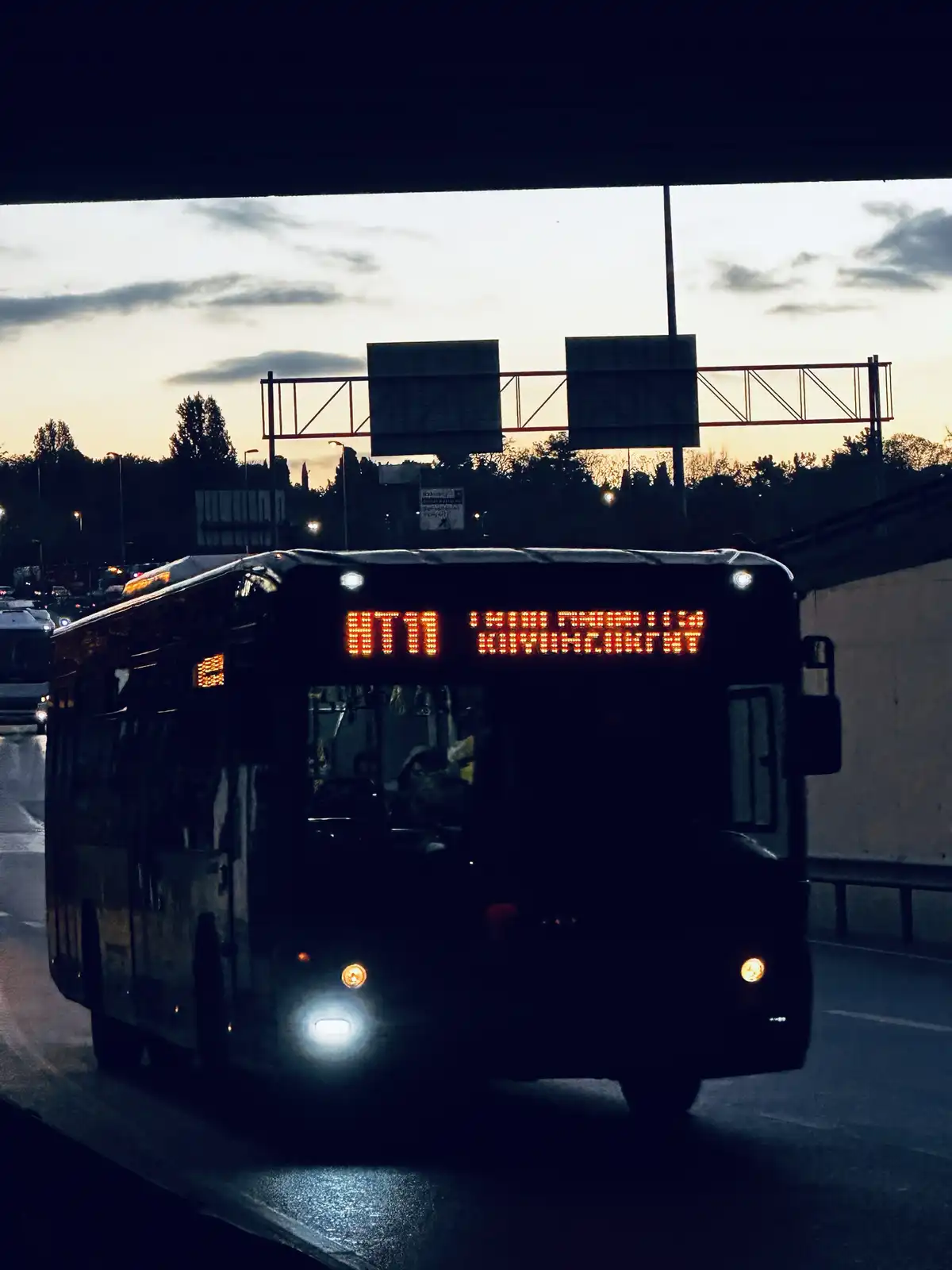 A dark bus is driving under an overpass at dusk, its front display reads 'ATM SALVATION' in orange LED letters. The scene is…