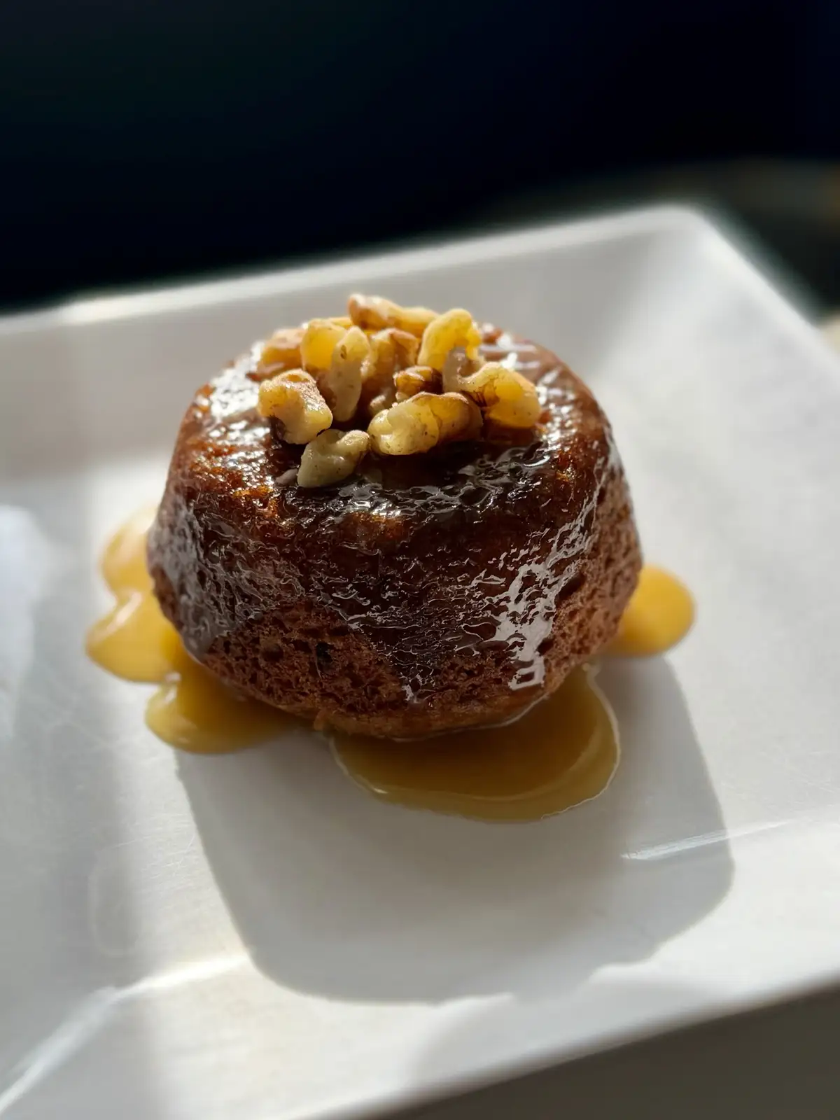 A close-up photo shows a small, round, caramelized dessert, likely a flan or pudding, topped with chopped nuts and drizzled…