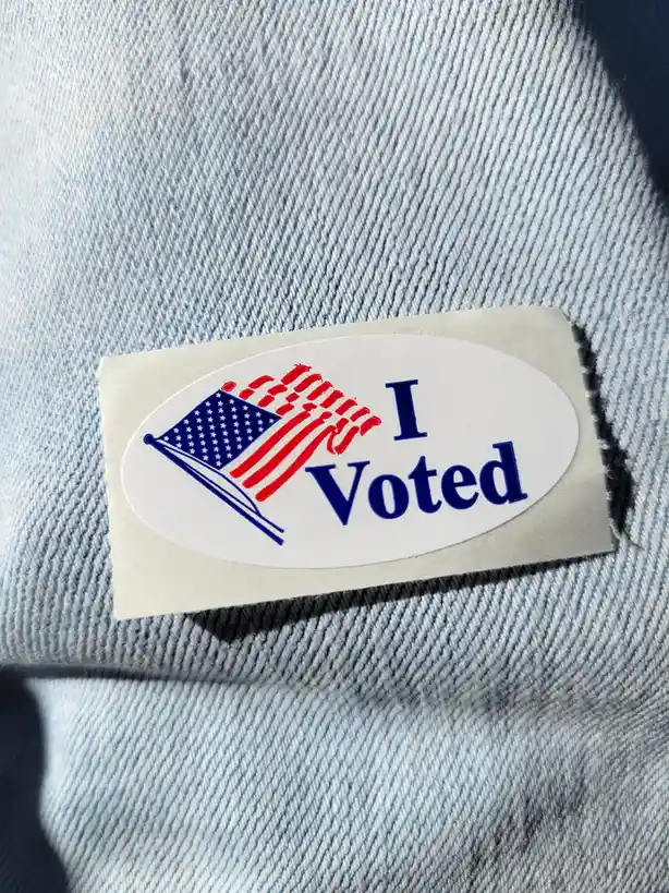 A close-up photo shows a white oval 'I Voted' sticker with a stylized American flag graphic, stuck on the denim fabric of a…