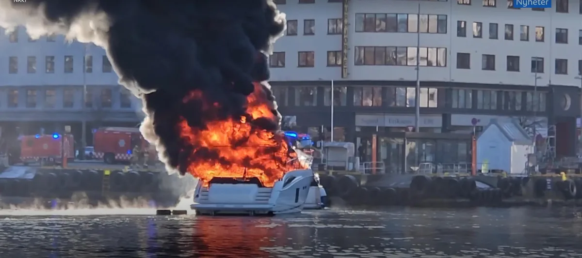 A white motorboat is engulfed in large flames and billowing black smoke while docked in a harbor, with emergency vehicles…