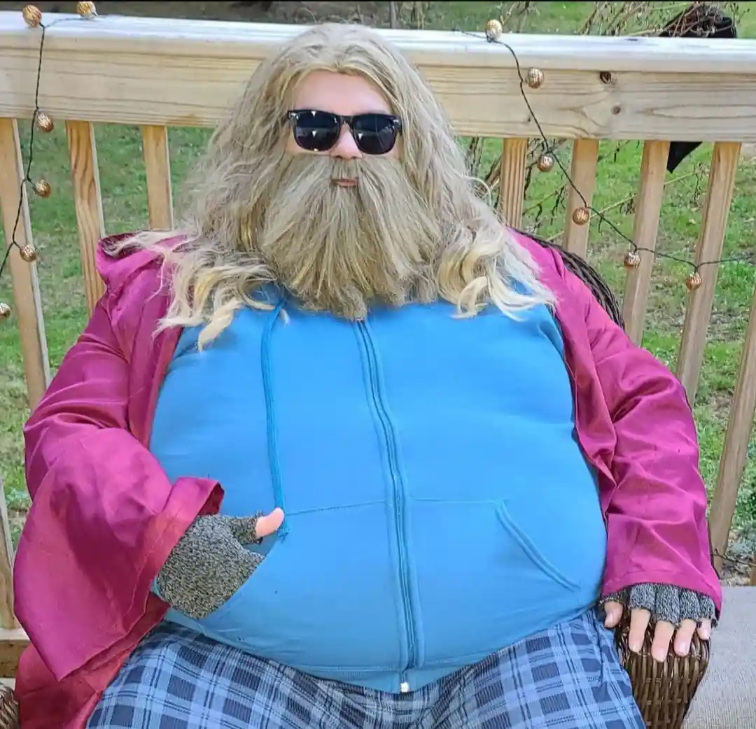 a person with a long blonde wig and beard, wearing sunglasses, sits on a wicker chair on a wooden deck. They are dressed in…