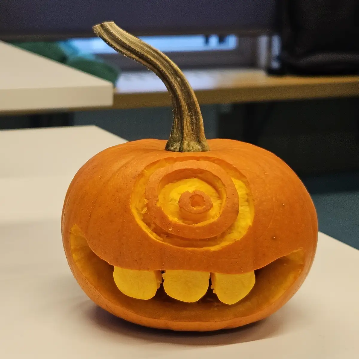 A small orange pumpkin sits on a white surface, carved with a spiral pattern for an eye and three rounded teeth in a mouth…