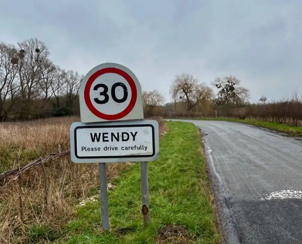A road sign on a rural road shows a standard 30 mph speed limit sign above a custom sign that reads 'WENDY Please drive…