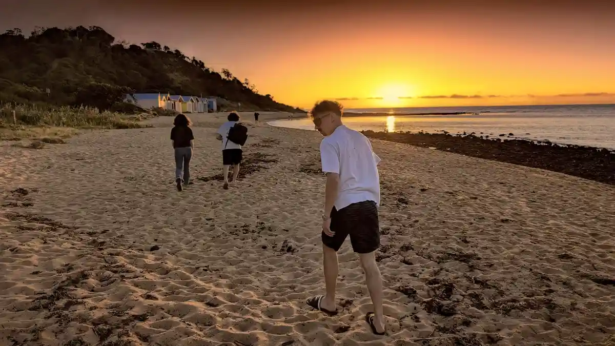 A man in a white t-shirt and dark shorts stands on a sandy beach at sunset, looking down at his feet while two other people…