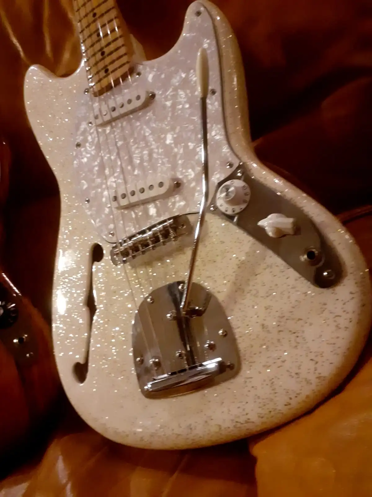 A white, speckled electric guitar with a microphone stand inserted vertically through its body, positioned near the bridge…