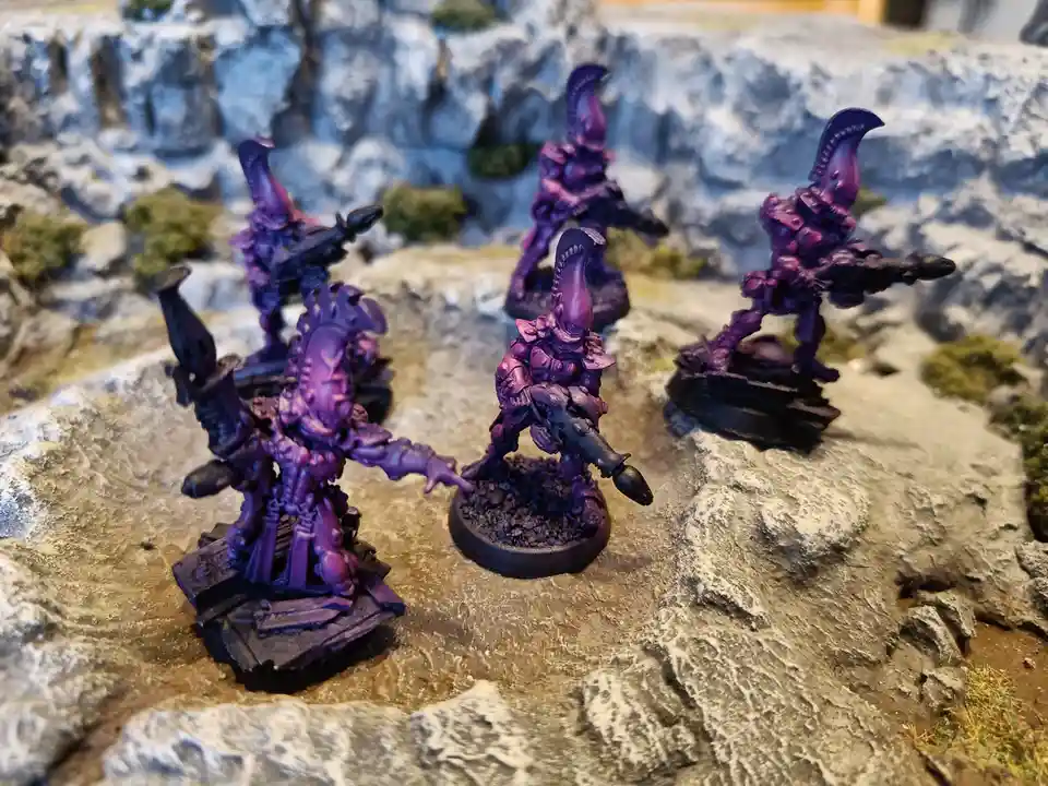 Five purple-painted miniature alien warriors with large curved horns and weapons stand on rocky terrain. One figure in the…