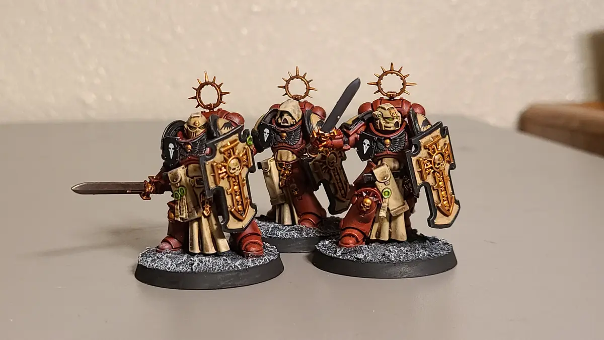 Three hand-painted miniature figurines of armored warriors, likely from Warhammer 40,000, stand on textured bases. Each…