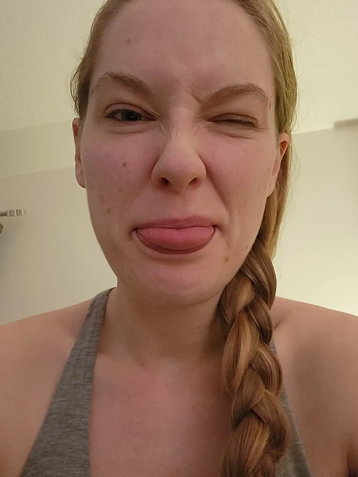 A close-up selfie of a woman with light brown hair in a braid, wearing a grey tank top, making a strong, exaggerated crying…