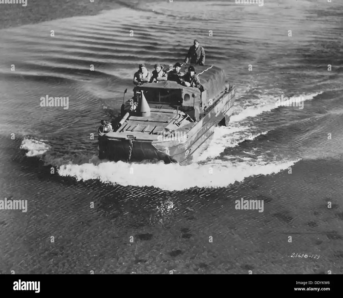 A black and white historical photo shows a military amphibious vehicle, resembling a DUKW, moving through water with a wake…