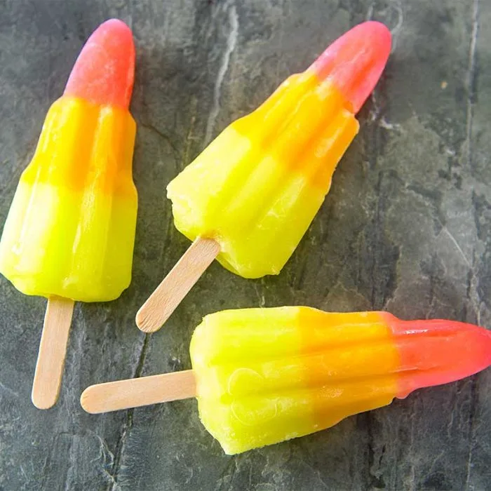 Three colorful, gradient-colored popsicles with wooden sticks lie on a dark, textured stone surface. The popsicles feature a…