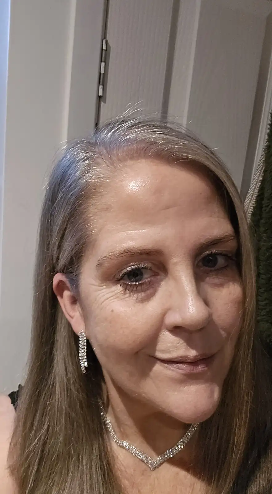 A close-up selfie of a woman with long, light brown hair showing gray roots, wearing sparkly diamond earrings and a matching…