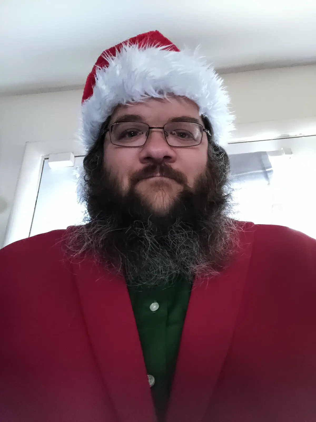 A man with a long, dark beard and glasses wears a red Santa hat and a red blazer over a green shirt, looking directly at the…