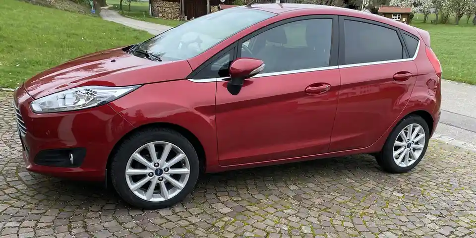 A red Ford Fiesta hatchback is parked on a cobblestone driveway in a grassy, rural setting. The car is shown in profile…