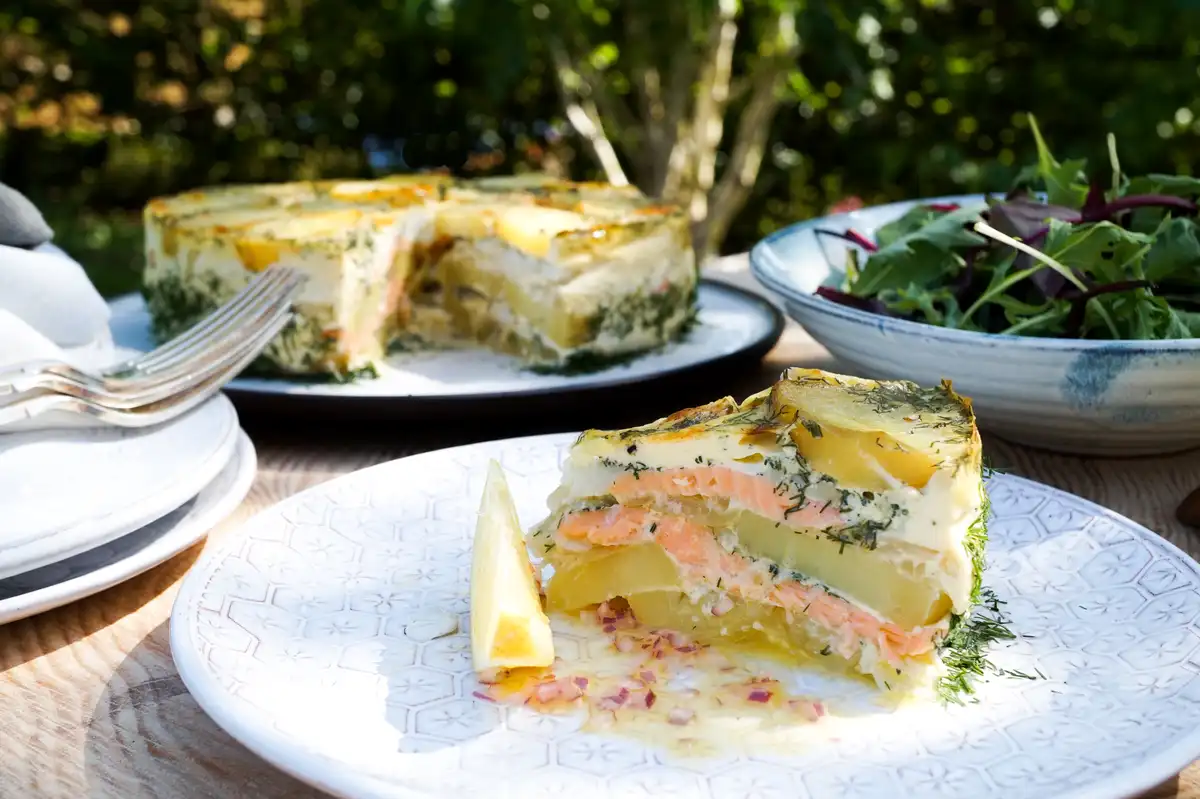 A slice of layered salmon and potato dish, resembling a cake, is served on a patterned plate with a lemon wedge and chopped…